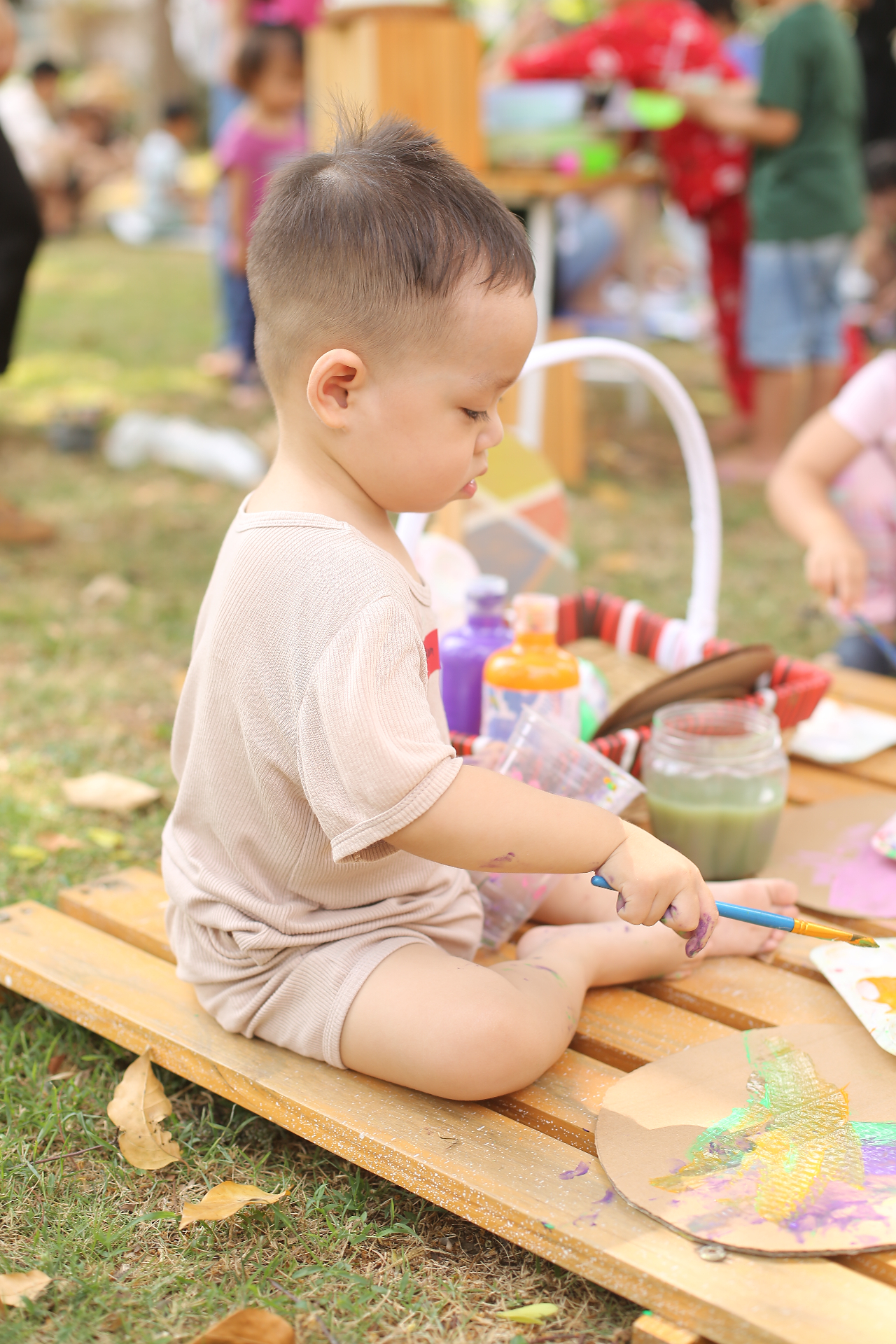 Bé trai tập trung cầm cọ vẽ trên bục gỗ ngoài sân — buổi messy play thật của CLB Lấm Lem
