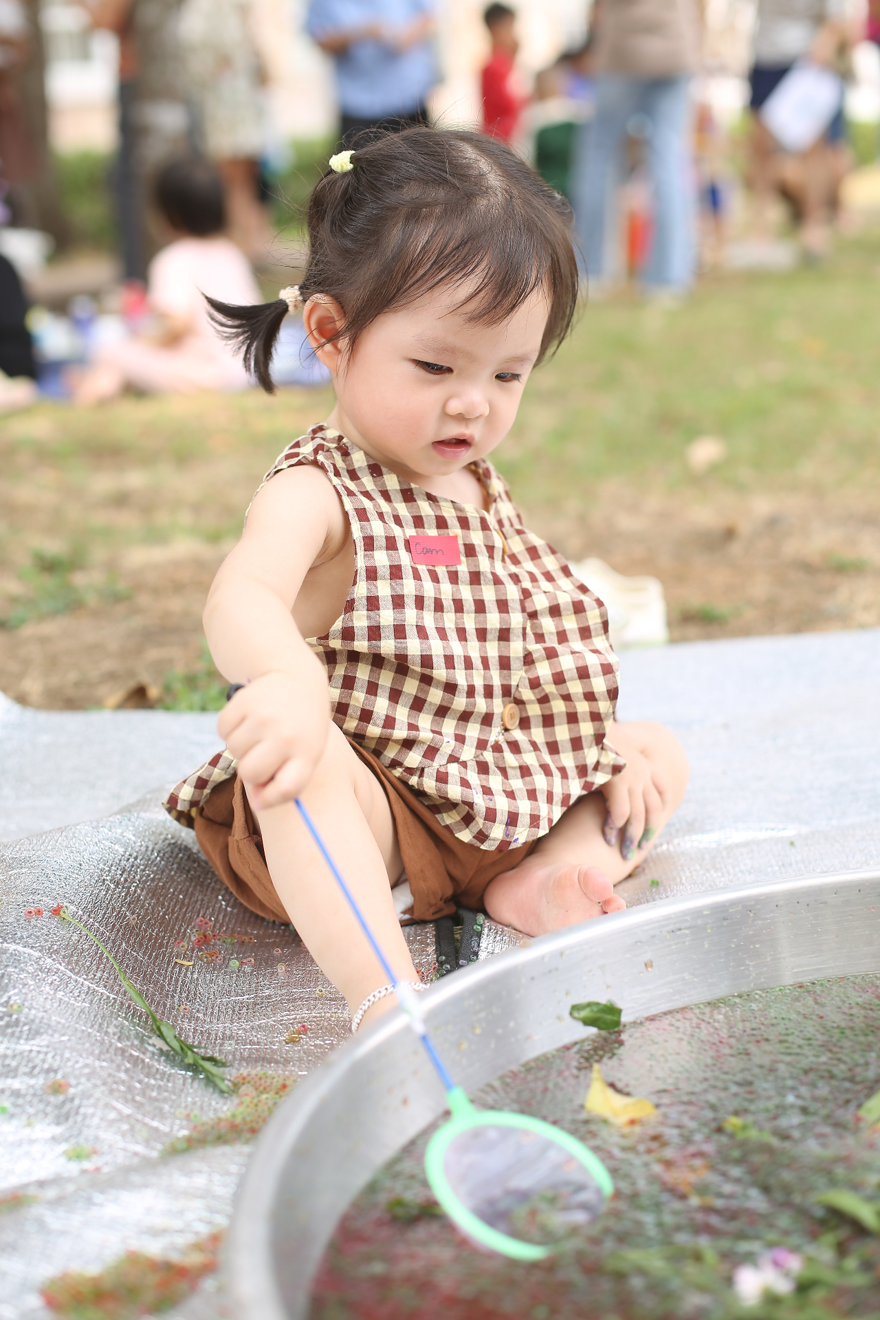Bé gái mặc yếm caro chơi với khay water beads ngoài trời tại buổi trải nghiệm Lấm Lem